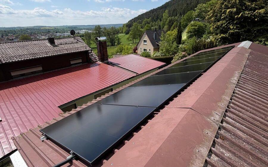 Acht schwarze Solarmodule auf einem roten Trapezblechdach in Kronach-Neuses, dahinter der Frankenwald