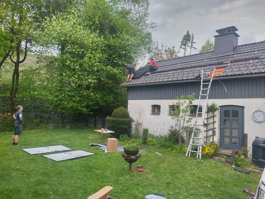 Zwei Monteure legen ein Solarmodul auf die Montageschienen eines Satteldachs, ein dritter steht im Garten, an der Traufe lehnt eine Leiter