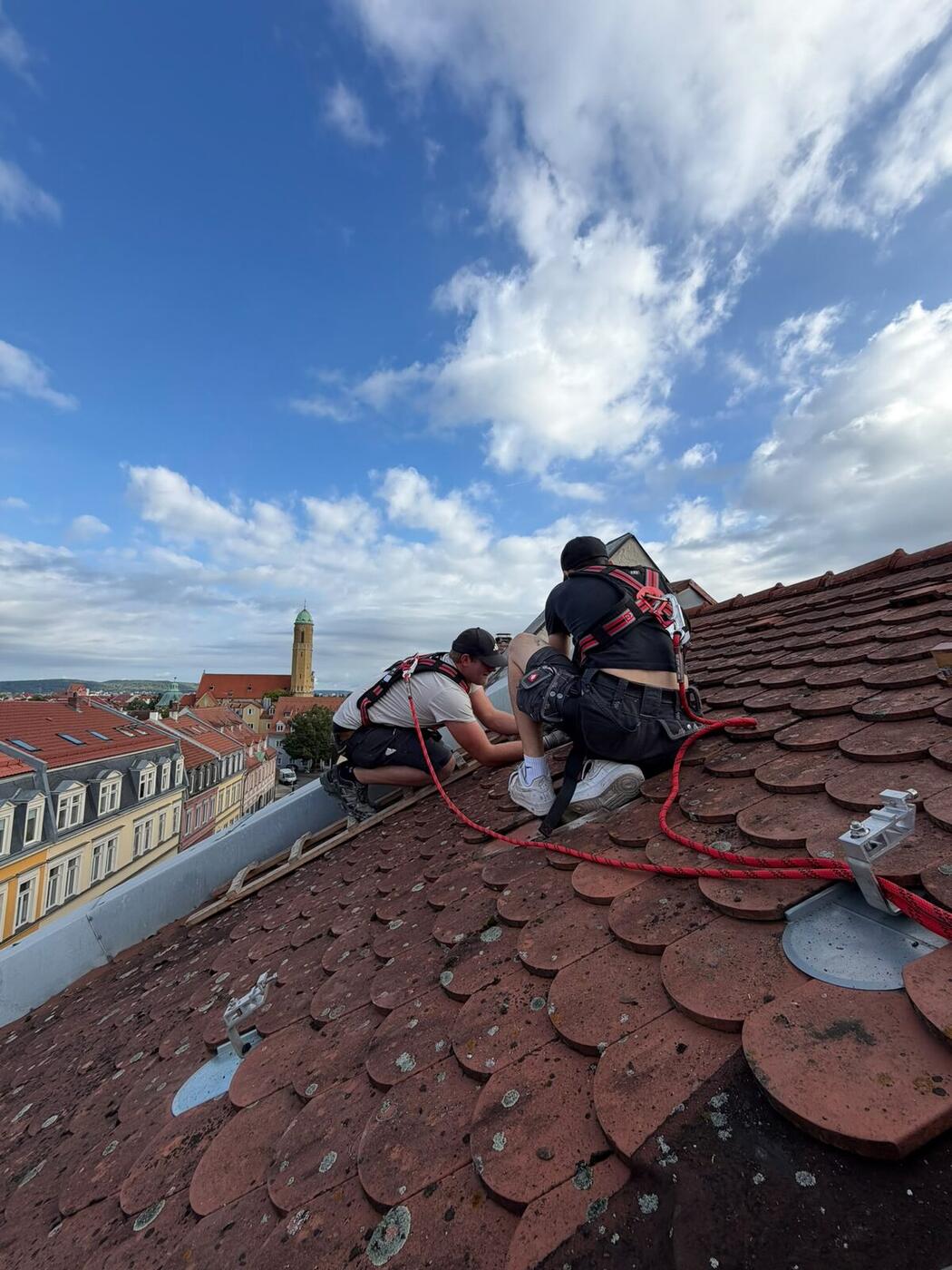 Zwei Monteure mit Auffanggurt und rotem Sicherungsseil setzen Dachhaken auf einem Biberschwanzdach in der Bamberger Altstadt