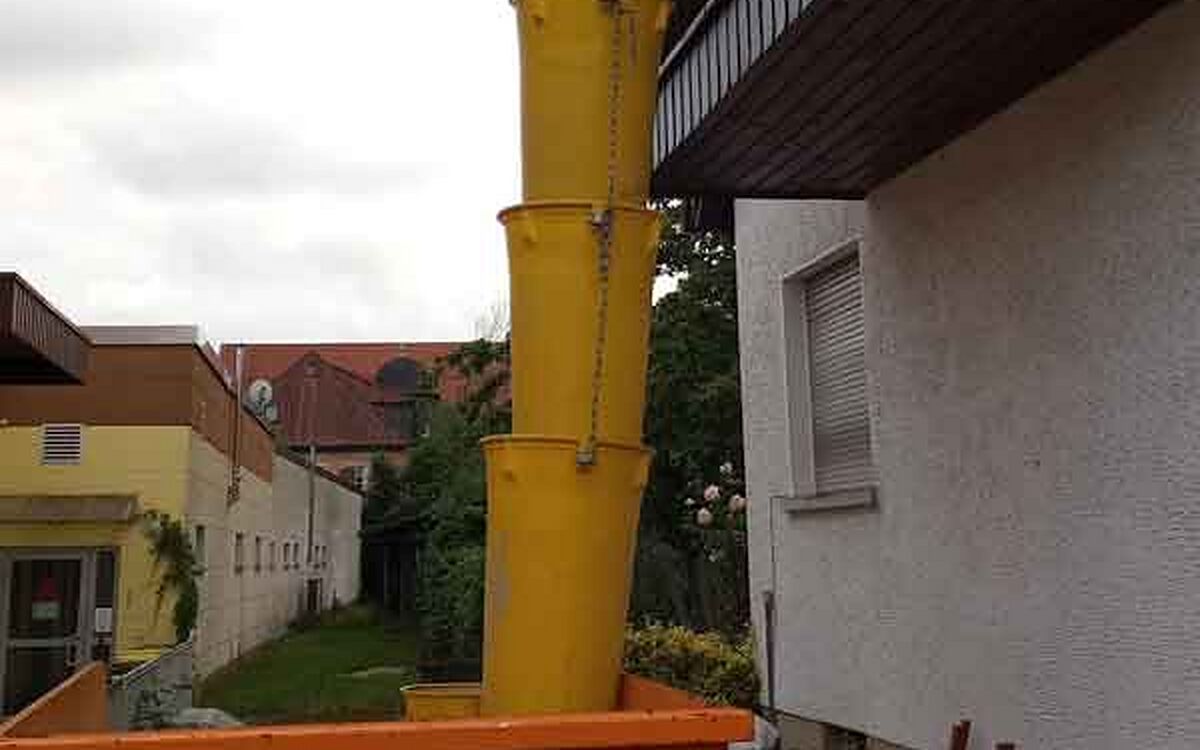 Gelbe Schuttrutsche am Balkon eines Wohnhauses, die Segmente führen hinunter in einen Container